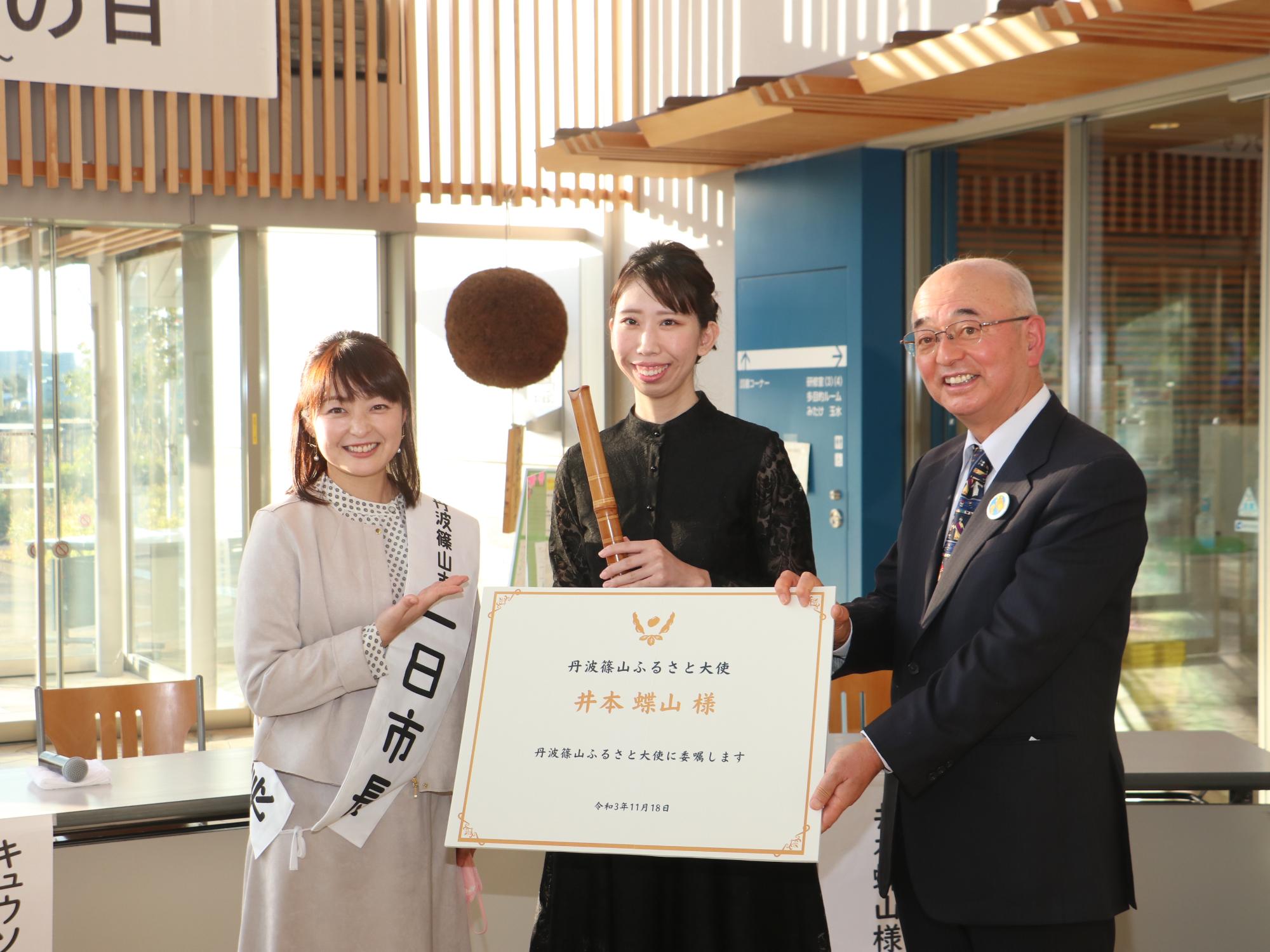 一日市長の熊谷さん、尺八を持った井本さん、井本さんへの大きな委嘱状を持った酒井市長との記念撮影