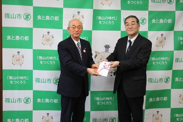 篠山市と書かれた壁の前でメガネをかけたスーツ姿の男性と目録と書かれた金封を受け取っている市長が並んでいる写真