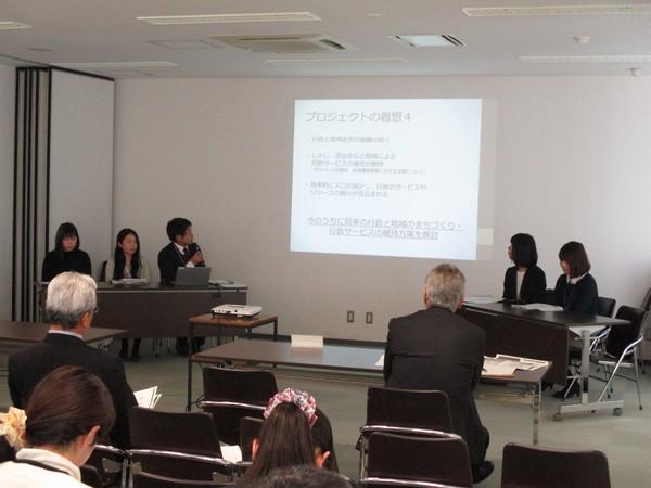 長机に職員5名が座っており、その中の男性職員がプロジェクターから映し出されている案件についてマイクで発表しているのを聞いている職員の写真