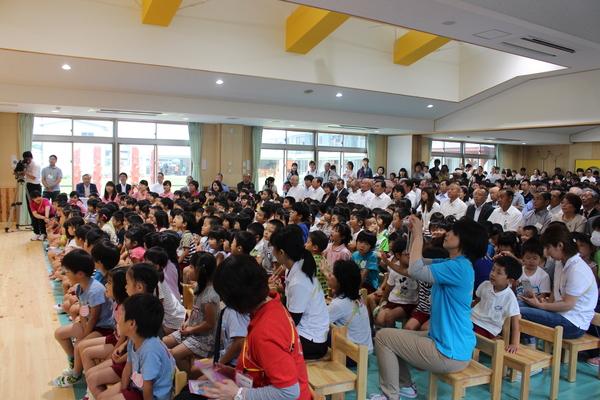 子ども園のホールの中で80名ほどの保育園生が木の椅子に座っていて、その後ろにスーツ姿の大人達が100名ほど座っている写真