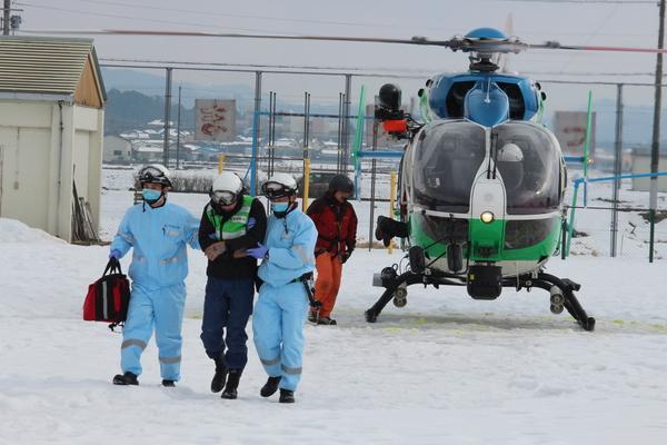 雪の降り積もっている中、ヘリコプターが停まっており、水色の防寒着を着た男性2名に青い防寒着を着た男性が両脇を抱えられ歩いている様子の写真