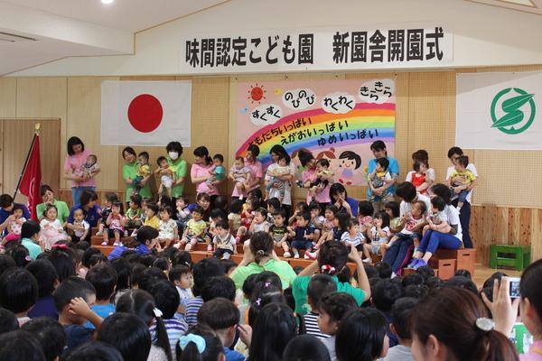 10名ほどの保育士が赤ちゃんを抱っこしていて立っていて、その前に30名ほどの保育園児が座っている写真