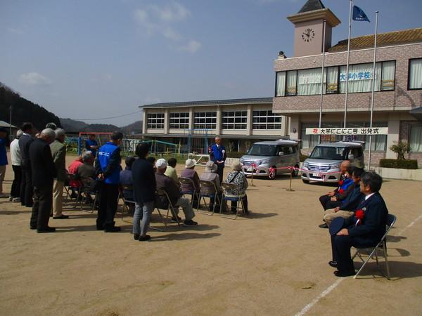 大芋小学校の運動場に出発式の会場が設置されており、グレーの自家用車2台が展示され、車の前には出発式の参加者が集まって式に参加している様子の写真