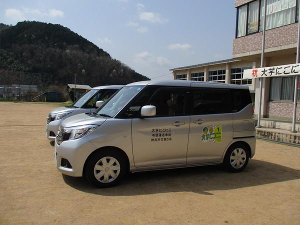 「大芋 にこにこ」という文字とお年寄りのイラストが描かれているグレーの自家用車2台を右横から写している写真