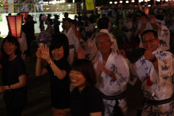 舞台下で踊りを踊っている笑顔の市長の肩に手をのせピースをしている眼鏡の男性とその前を歩く3人の笑顔の女性の写真