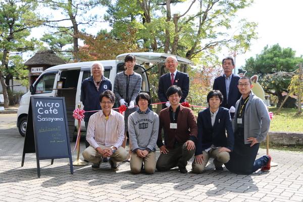 移動カフェの車の前で5名の男子大学生と、市長、職員が記念撮影をしている写真