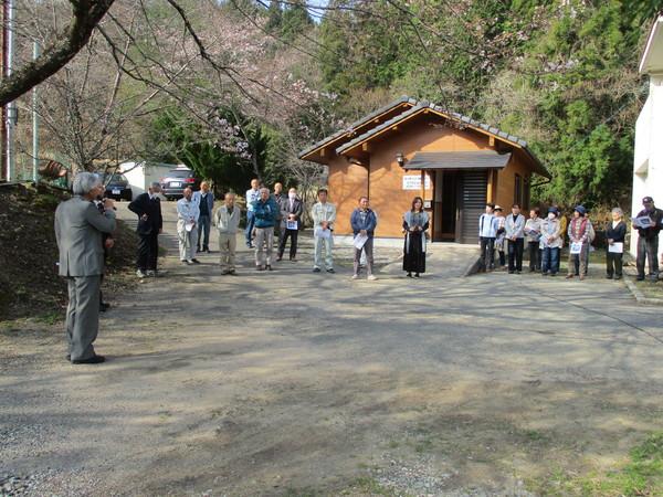 さくらの木の下に出発式に参加している人が資料を手に持って集まっており、マイクを持っている白髪の男性が話をしている様子の写真