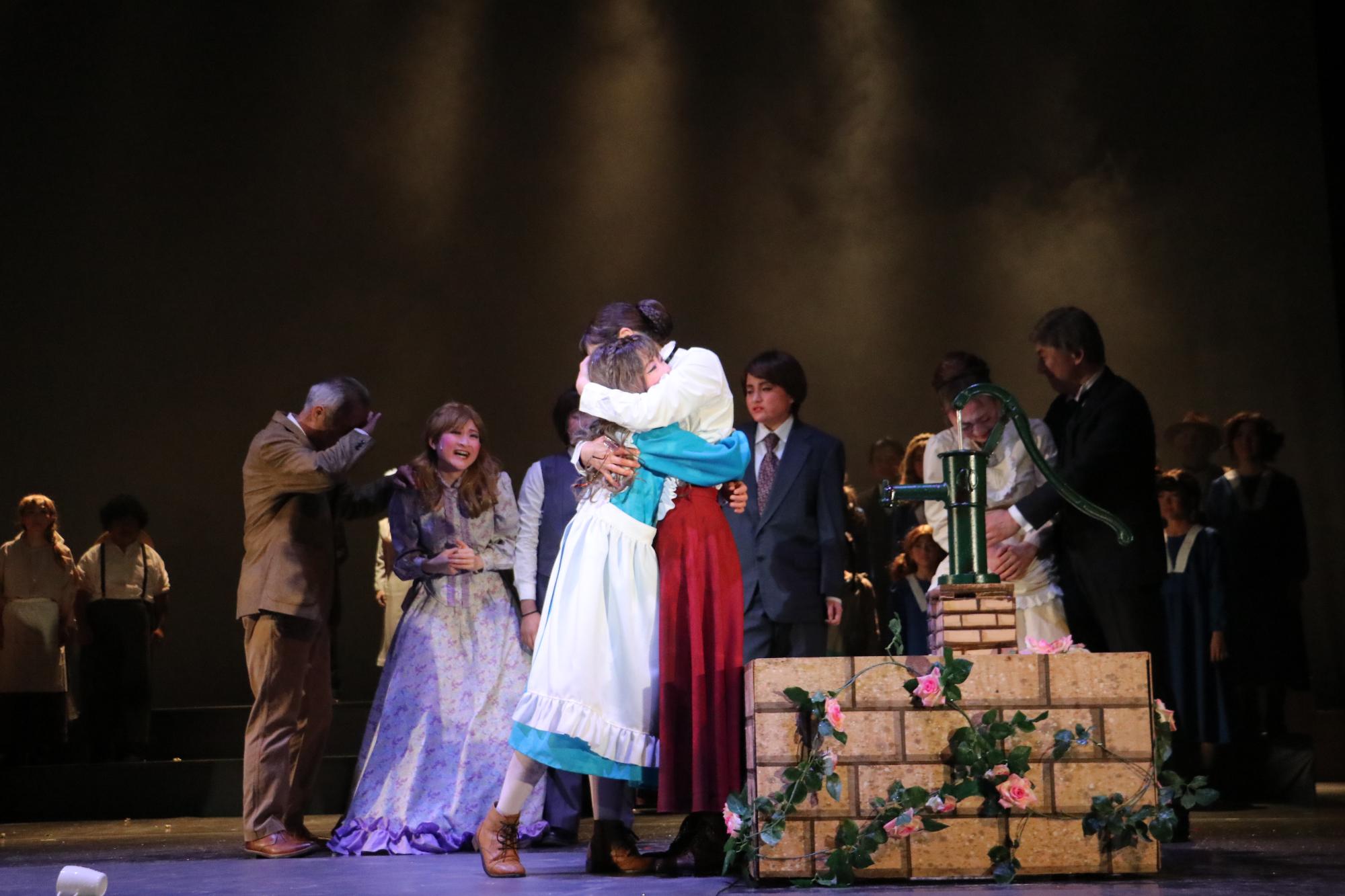 井戸のようなところのそばで、ヘレンとサリバン先生がお互いに抱きしめ合っていて、その後ろで数人の大人たちがその姿を見つめている写真