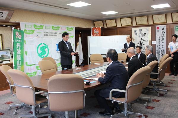 応接室の会議テーブルに座る市長とスーツを着た男性の前で立って紙を読み上げる男性の写真