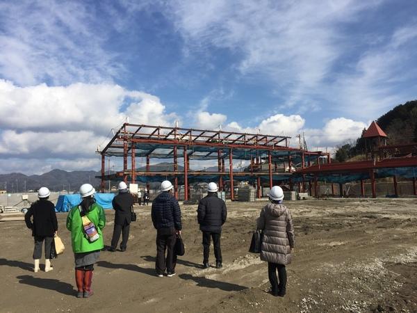 青空の下に建設中の鉄筋の骨組みがあり、それをヘルメットをかぶった6名の人達が見ている写真