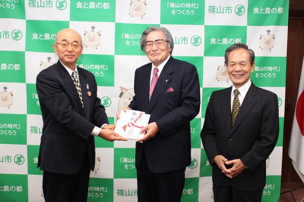 村岡 万巧代表取締役社長が市長へ寄付金を手渡しで渡しており、犬伏 照人顧問と3人で写っている写真