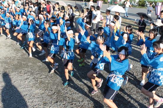 祭りの法被を着た子供達が両手を右上にあげ踊っている様子を上からうつした写真