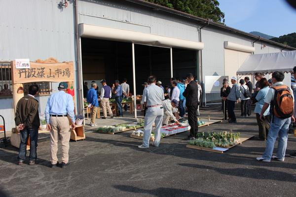奥には倉庫があり、その手前にきゃべつ、トマトなどの野菜が置かれ30名ほどの来場者達が立っている写真