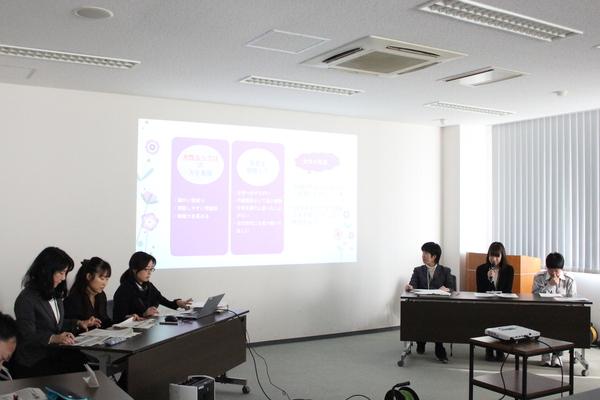 長机に女性職員6名が座っており、その中の職員がプロジェクターから映し出されている案件についてマイクで発表している写真