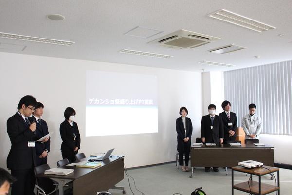 プロジェクターから映し出されている「デカンショ祭盛り上げプロジェクト提案」の発表で担当チーム7名が立っており、その中の男性職員がマイクで発表している写真