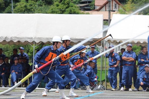 番号ゼッケンをつけた消防団員3名がホースと鎌のようなものを持ち放水している写真