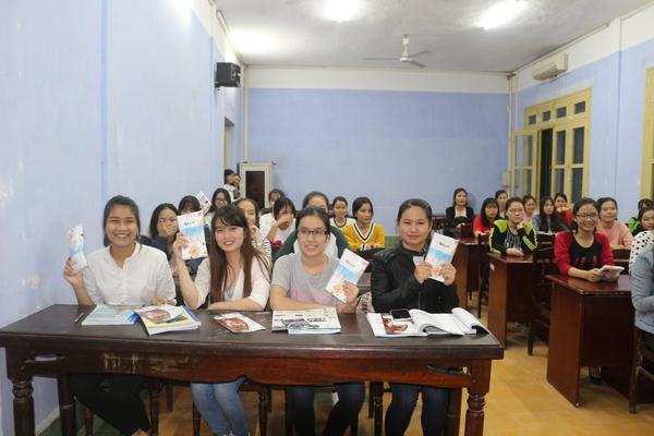 20名以上の女子学生たちが座っていて、一番前にいる4名の学生たちが手に教科書をもって微笑んでいる写真