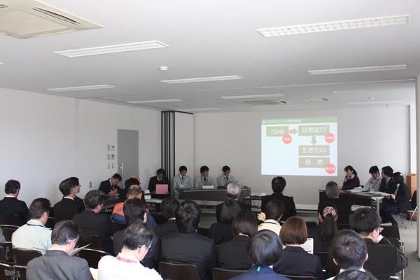 長机に職員8名が座っており、その中の女性職員がプロジェクターから映し出されている案件についてマイクで発表しているのを聞いている職員の写真