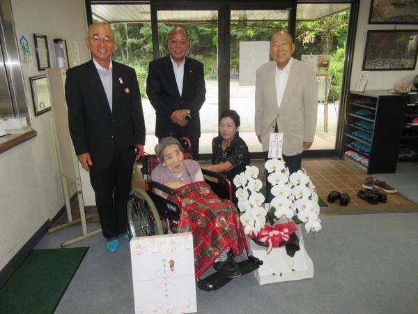 車いすに乗った瀬々倉きみ江さん(101歳)がエントランスで市長らとお祝いの胡蝶蘭と記念品を前に記念撮影している写真