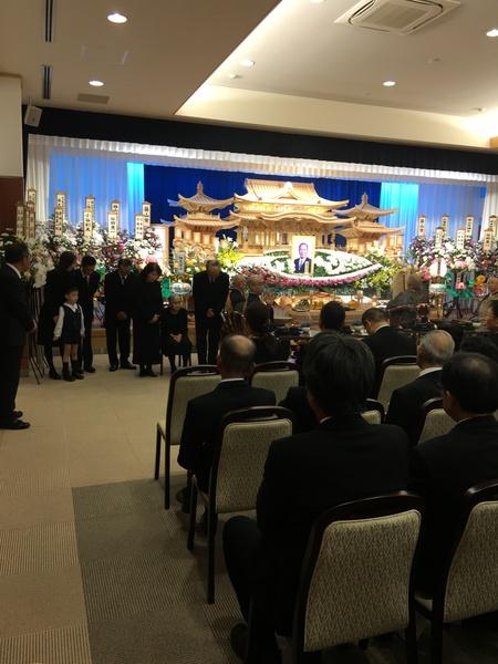 沢山のお花に囲まれた祭壇と祭壇の前に座っているお坊さんと立っている親族の方々と椅子に腰かけている参列者の写真