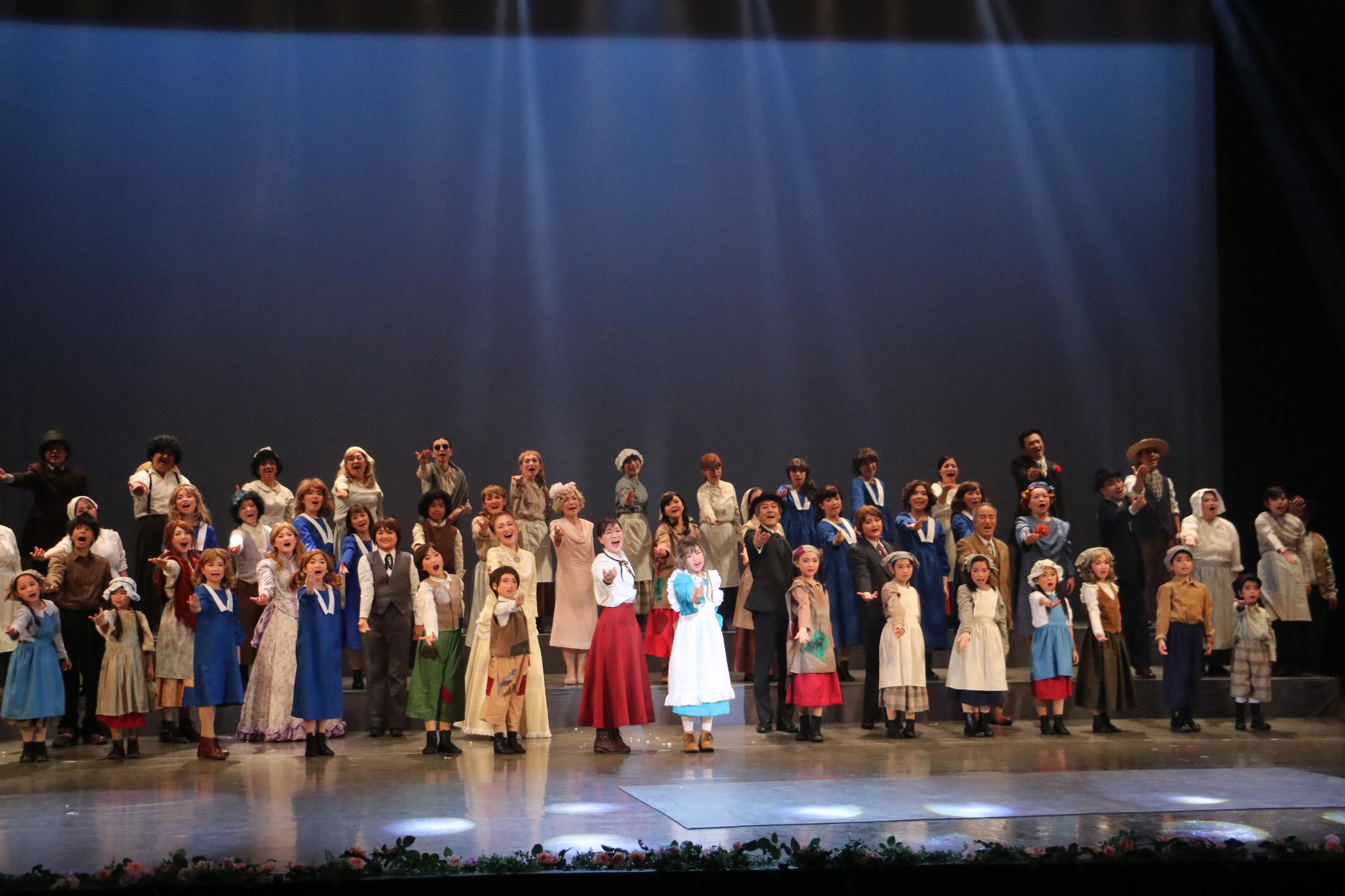 真ん中にヘレンとサリバン先生、その横にはたくさんの子どもたち、後ろには大人の出演者の皆さんが右手を横にあげ歌っている写真