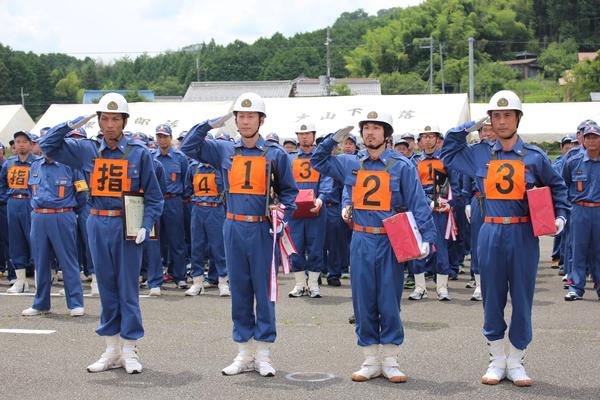 整列する消防団員の前で4名の団員が賞状と表彰盾と景品を持ち敬礼している写真