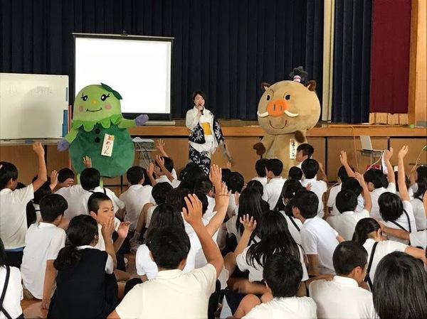 小林 真弓さんが篠山市のマスコットキャラクターと一緒に子供達の前に立ち、子供達が手を上げて質問している写真