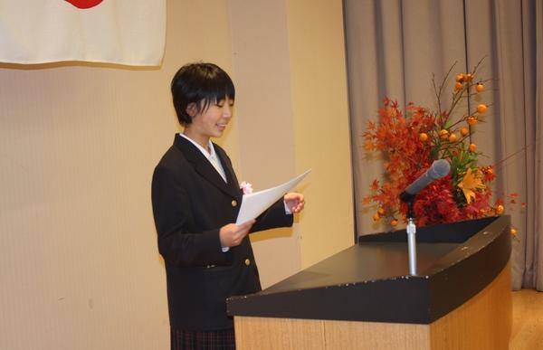 壇上花の置かれた演台の前で、原稿のようなものを持って立っている小林 優花さんの写真