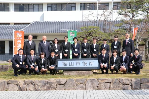 外にある篠山市役所と書かれた石碑を囲んで記念撮影をしている新規職員と市長、関係職員の写真