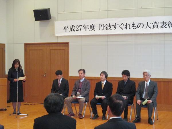 表彰式で司会者の女性が発表している写真