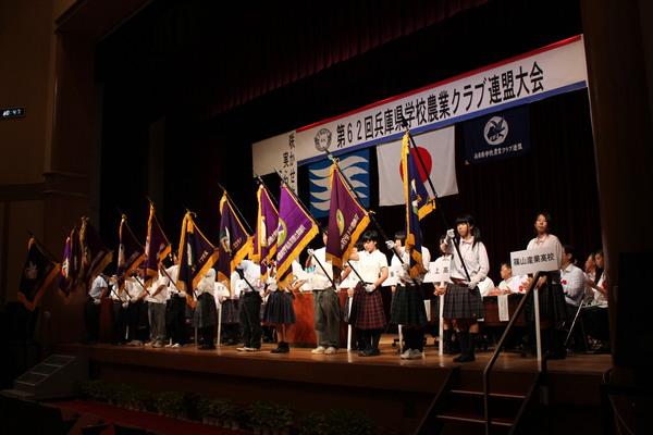 各高校の生徒の代表が壇上に上がり、校旗を持って立っている様子の写真