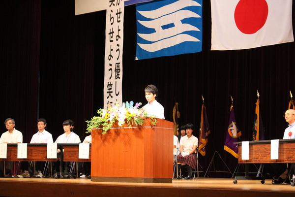 壇上でクラブ連盟会長の山口君が挨拶をし、後方には関係者が椅子に座って話を聞いている様子の写真