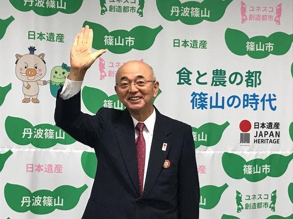 市長が篠山市と書かれた壁の前で右手を上にあげて、笑っている写真