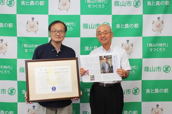 受賞された賞状の入った額をもっている山中さんと、山中さんの記事が載っている雑誌を広げて持っている市長が並んで写っている写真