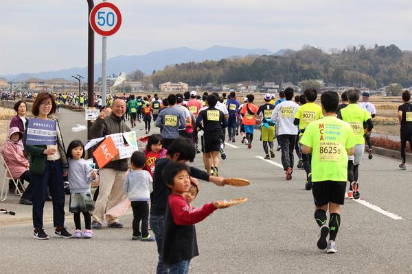 走っているランナーを応援している子供2人がザルに入っているチョコレートを差し出している写真
