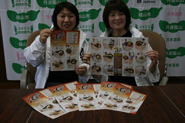 白いパーカーを着ている左側の女性は「篠山まるごと丼」のパンフレットの表紙を、右側の女性は広げて両手に持っている写真