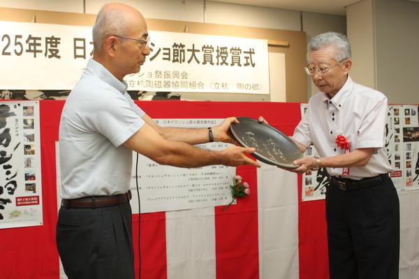 胸章リボンをつけた酒井 修さんが、市長からまるいプレート受け取っている写真