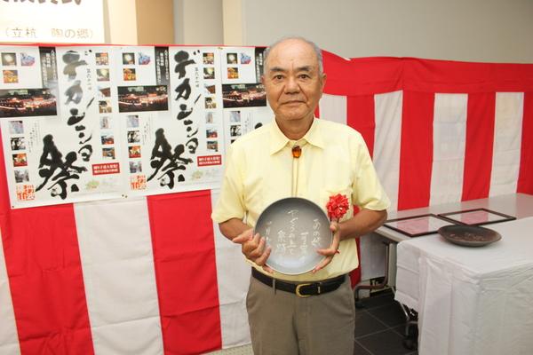 胸章リボンをつけ黄色いシャツを着た辻田 積男さんが、まるいプレートを持っている写真