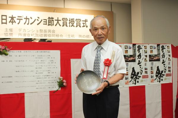 胸章リボンをつけスーツを着た佐古田 正明さんが、まるいプレートを持っている写真