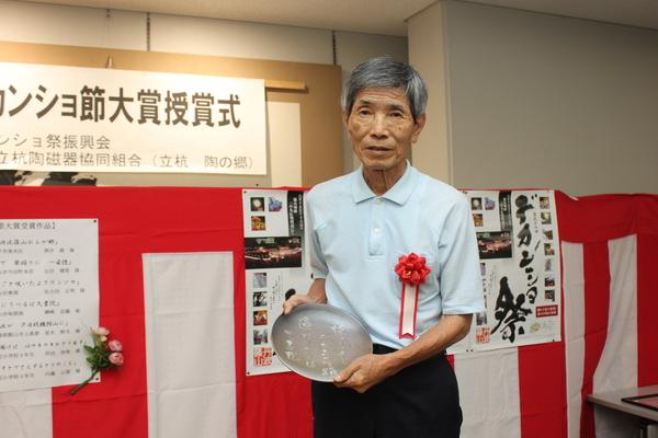 水色のポロシャツに胸章リボンをつけた御崎 忠義さんが、まるいプレートを持っている写真