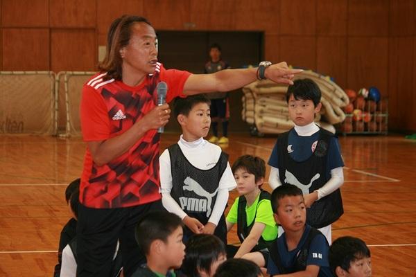 体育館でマイクを持った北澤選手が指をさして子供たちに話をしている写真