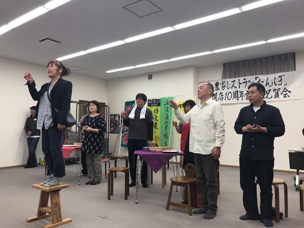 コップを手に持った女性が椅子の上に立っていて、その後ろの人達もコップを持って上に掲げている写真