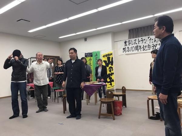 4名の男性が立っていて、奥にいる白い服の男性が、手前にいる男性を指さしながら話をしている写真