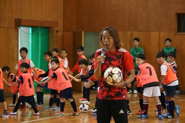 体育館でボールを片手に持った北澤選手がマイクで話しているのをビブスをつけた子供たちが手を繋いで見ている写真