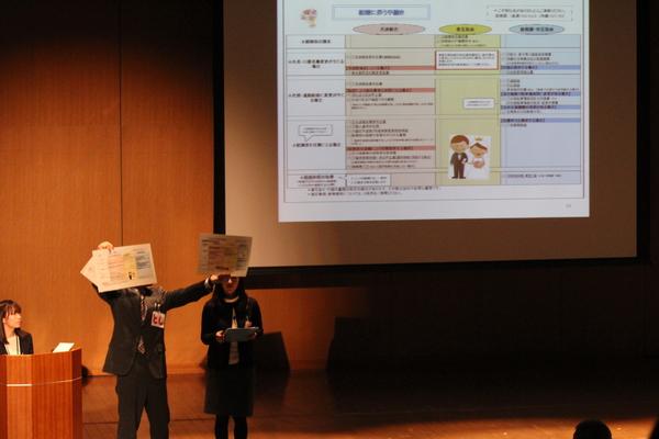 舞台にてスクリーンに映し出された案件について発表する職員の写真