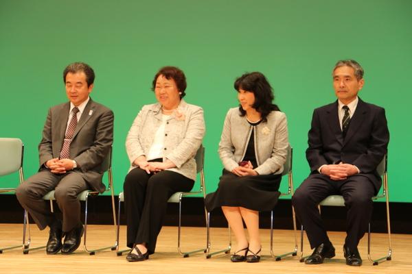 後ろの壁紙は緑色で、スーツを着た男性2人、女性2人が椅子座っている写真