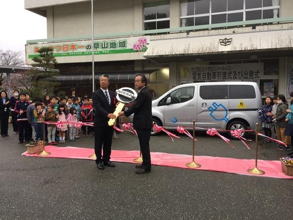 建物の所にワンボックスカーが置いてあり大きな手作りの鍵を受け取っている写真