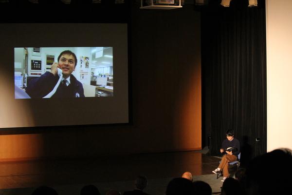 舞台のスクリーンに映し出された電話に出ている男性の映像について、椅子に座った男性職員が説明をしている写真