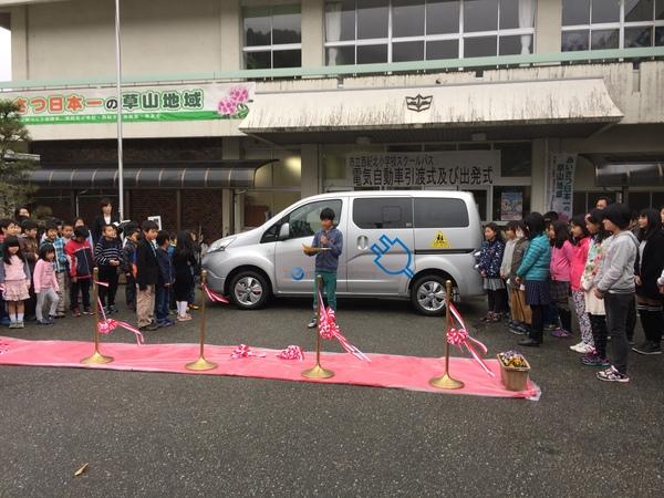 ワンボックスカーの前で乗本君が紙を手に持ちマイクでお話ししている写真