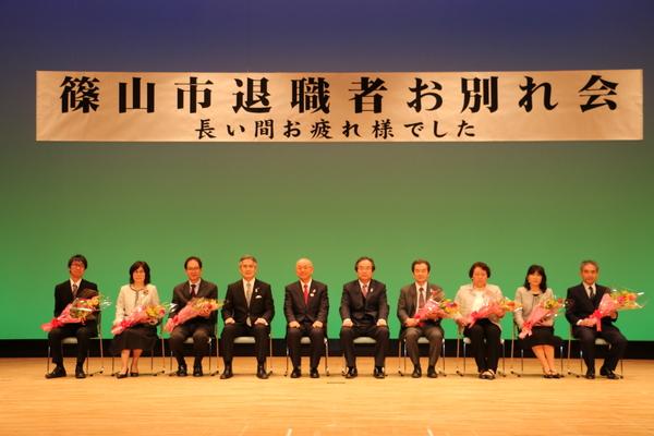 市長とスーツを着た方が9名座っていて、右4人と左3名が花束を持って記念写真